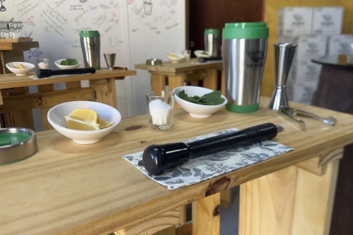 Wooden tables with lemons, mint, glasses, shakers, and a muddler in a cozy room.